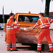 Zwei Arbeiter in Warnkleidung laden Erde auf ein Fahrzeug.
