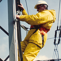 Ein Mann arbeitet an einem Strommast, er tägt einen wetterfesten Anzug.